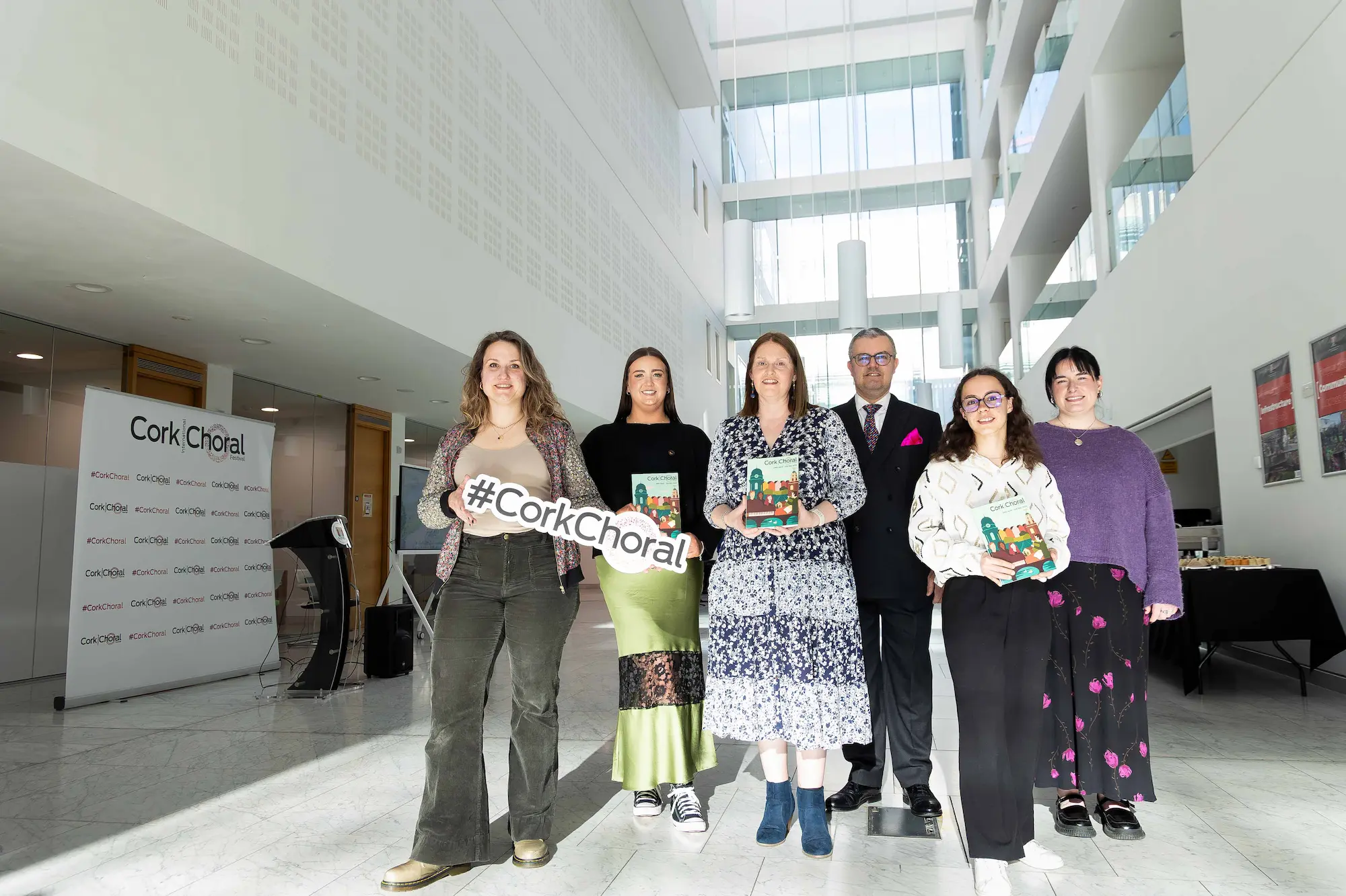 Cork Choral Festival team posing together in a bright venue, with members holding Cork Choral Festival programmes and a #CorkChoral sign.